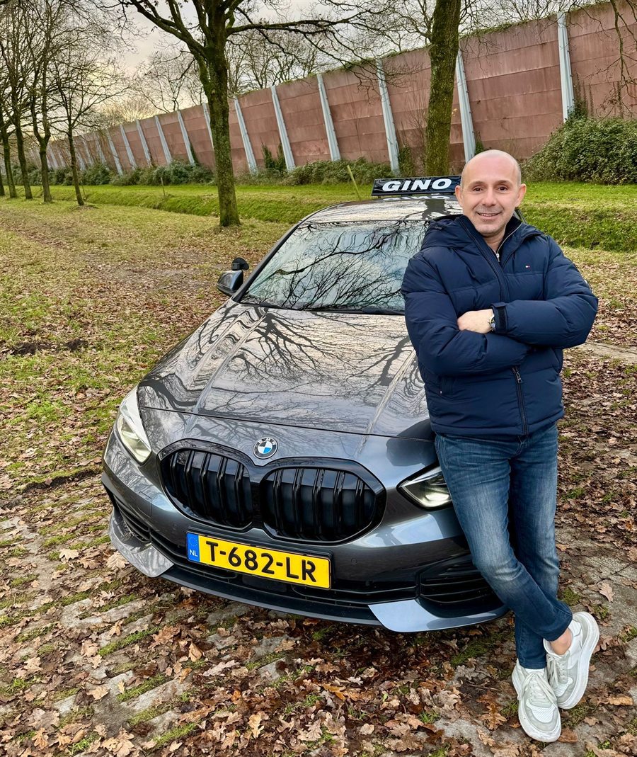 Gino Peshdary bij zijn grijze BMW-lesauto met GINO-daklichtbak